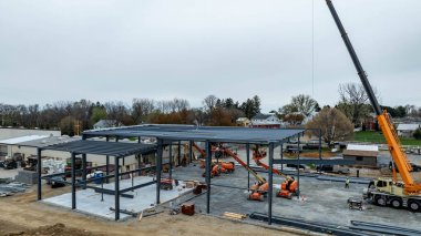 Yeni bir binanın inşa edildiği inşaat sahasında çalışmalar sürüyor. İşçiler ve makineler aktifken vinçler çelik kirişleri kaldırır. Hava bulutlu, yılın hangi zamanı olduğunu gösteriyor..