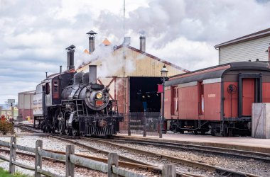 Bir buharlı tren tren tren garının yanındaki istasyona yuvarlanıyor. Motor yaklaşırken dumanlar yükseliyor. İnsanlar yakınlarda gelişi izliyor olabilir. Atmosfer hareketli ve yoğun..