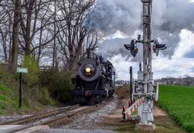 Strasburg, Pennsylvania, 6 Nisan 2024 - Demiryolu kavşağının yakınından bir buhar treni geçiyor. Gökyüzü bulutlu ve manzaranın yeşil tarlaları ve ağaçları var. Tren duman üretir.