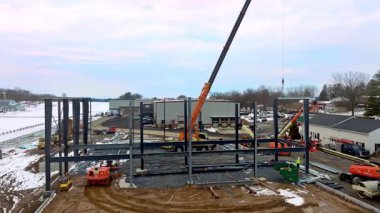 Gordonville, Pennsylvania, 25 Şubat 2026 - Büyük vinçler yeni bir bina için çelik kirişleri yerleştiriyor. İşçiler şantiyede makineler çalıştırıyorlar. Kar zemini kaplıyor, bu da kışı gösterir.