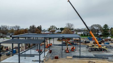 İşçiler gün içinde banliyö bölgesinde metal bir bina inşa ediyorlar. Bölgede birçok araç ve araç varken vinç malzemeleri kaldırır.