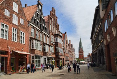 Luneburg, Almanya - 4 Temmuz 2019: İnsanlar tarihi binalar ve 4 Temmuz 2019 'da Almanya' nın Luneburg kentindeki St. Johannis kilise kulesiyle bir alışveriş caddesinde yürüyorlar.
