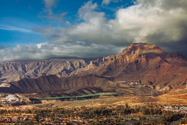 İspanya 'nın güneyindeki Tenerife, Kanarya Adaları' ndaki Montana de Guaza volkanik dağının dramatik manzarası. Engebeli uçurumlarla, doğal anıtlarla ve bulutlu gökyüzünün altında sıcak altın ışıkla kaplı çorak bir manzara. Manzaranın arka planı, seyahat ve turizm beldesi
