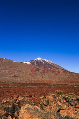 Teide Dağı 'nın muhteşem manzarası Teide Ulusal Parkı, Tenerife, Kanarya Adaları, İspanya' daki kırmızı volkanik lav sahasının üzerinde yükseliyor. Renkli kayalar, lav oluşumları ve berrak mavi gökyüzü ile dramatik volkanik manzara en büyük