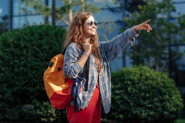 Sonriente joven rubia hipster mujer estudiante con mochila apuntando dedo lejos en la universidad. — Foto de Stock