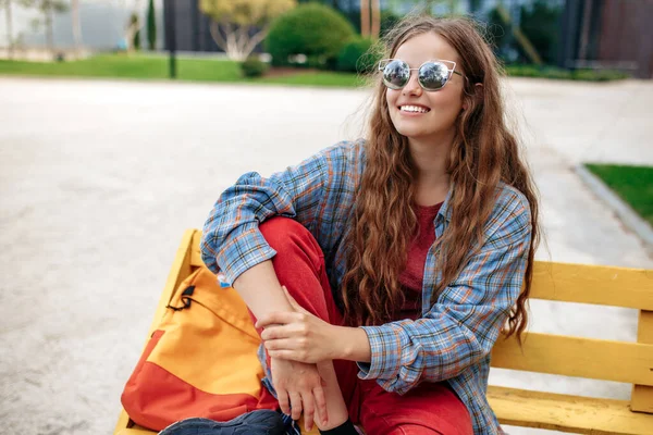 Glückliche junge blonde Hipster-Studentin mit Rucksack auf Bank an der Universität sitzend. — Stockfoto