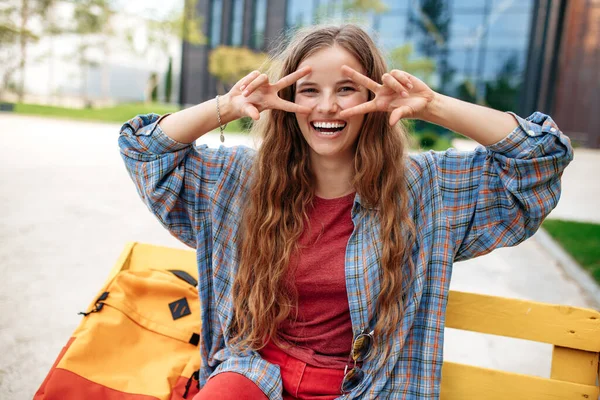 Cheerful young blonde hipster woman student with backpack looking to camera with happy face. Peace gesture. — Stock Photo, Image