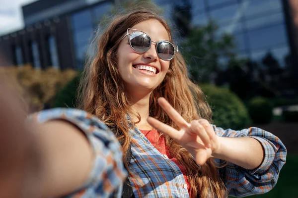 Feliz joven rubia hipster mujer estudiante con mochila tomar sefie con teléfono inteligente en la universidad. Gestos de paz. — Foto de Stock