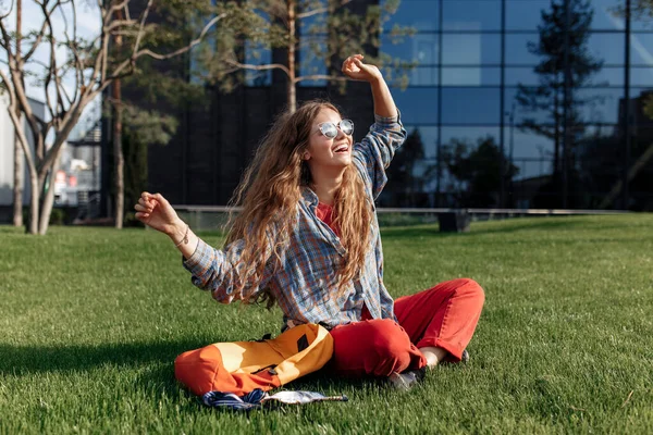 Alegre joven rubia hipster mujer estudiante con mochila sentado en la hierba en la universidad. — Foto de Stock