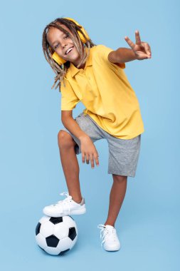 Bir bacağı futbol kulaklıklı, Afrikalı neşeli küçük bir çocuk mavi arka planda barış jesti topu gösteriyor..