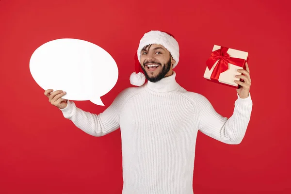 Feliz hipster homem usa santa claus chapéu segurando caixa de presente e fala bolha sobre fundo vermelho. — Fotografia de Stock