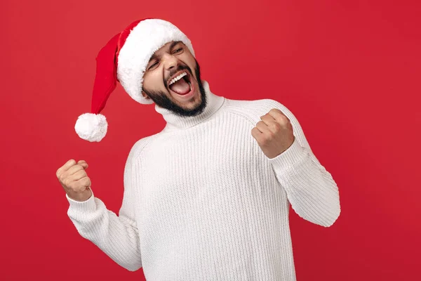 Spännande hipster man bär Santa claus hatt fira framgång över röd bakgrund. Jul- och nyårskoncept. — Stockfoto