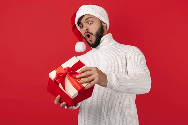 Homem hipster chocado usa chapéu de Papai Noel caixa de presente de abertura sobre fundo vermelho. Conceito de Natal e Ano Novo. — Fotografia de Stock