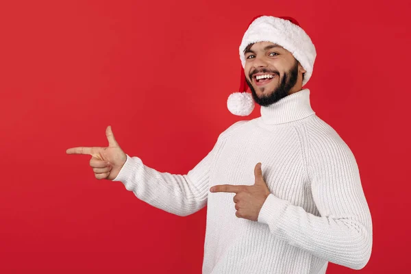 Bonito hipster homem usa chapéu de Papai Noel apontando dedos de distância sobre fundo vermelho. Conceito de Natal e Ano Novo. — Fotografia de Stock