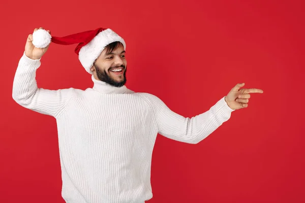 Homem hipster alegre usa chapéu de Papai Noel apontando o dedo para o fundo vermelho. Conceito de Natal e Ano Novo. — Fotografia de Stock