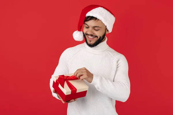 Homem hipster incrível usa chapéu de Papai Noel caixa de presente de abertura sobre fundo vermelho. Conceito de Natal e Ano Novo. — Fotografia de Stock
