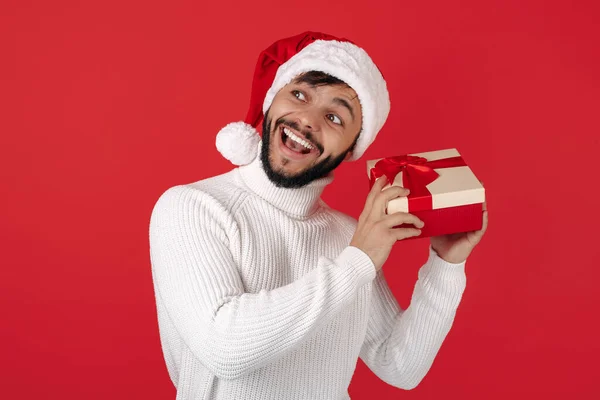 Förvånad hipster man bär Santa claus hatt hålla presentförpackning över röd bakgrund. Jul- och nyårskoncept. — Stockfoto