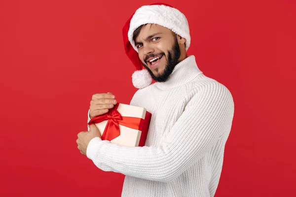 Hesitating hipster man bär Santa claus hatt hålla presentförpackning över röd bakgrund. Jul- och nyårskoncept. — Stockfoto