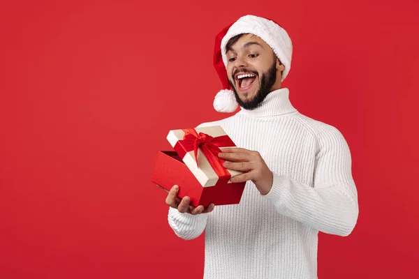 Homem hipster encantado usa caixa de presente de abertura de chapéu de Papai Noel sobre fundo vermelho. Conceito de Natal e Ano Novo. — Fotografia de Stock