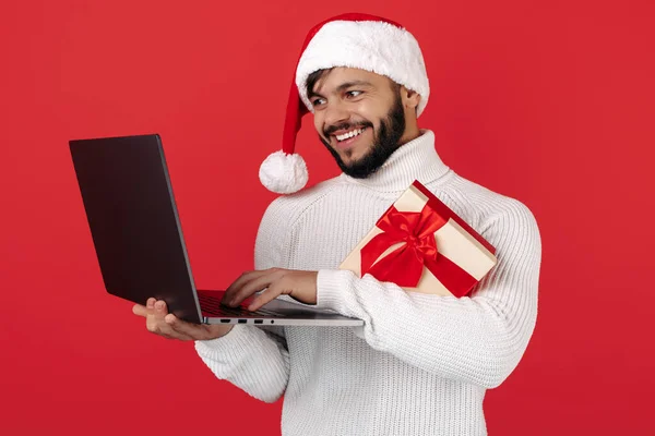 Homem hipster feliz usa chapéu de Papai Noel segurando notebook e caixa de presente sobre fundo vermelho. Conceito de Natal. — Fotografia de Stock