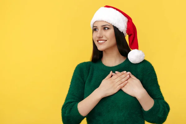 Söt ung brunett kvinna bära Santa claus hatt och grön pullover hålla händerna på bröstet över gul bakgrund. — Stockfoto