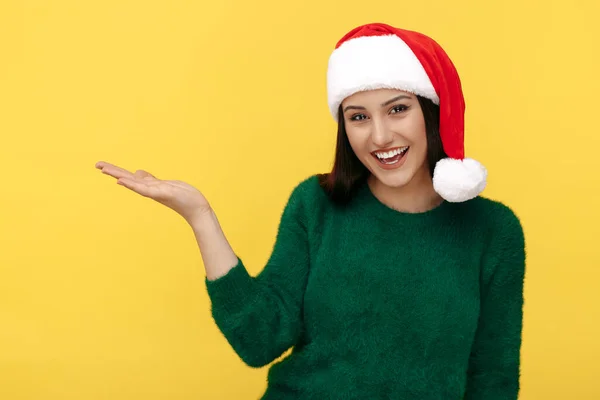 Glada unga brunett kvinna bära Santa claus hatt och grön pullover över gul bakgrund. — Stockfoto