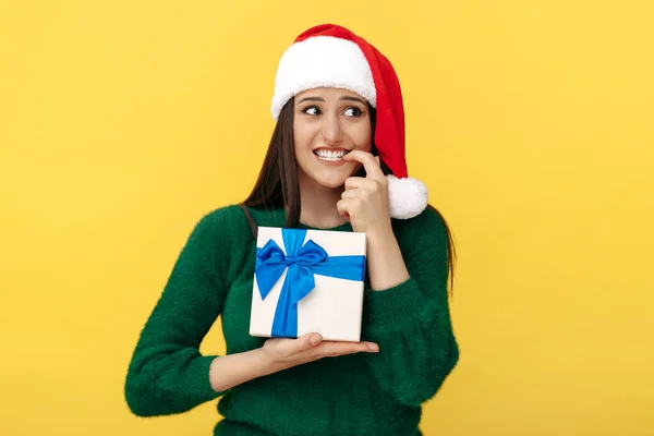 Hesitating ung brunett kvinna bära Santa claus hatt och grön pullover innehav presentförpackning över gul bakgrund. — Stockfoto