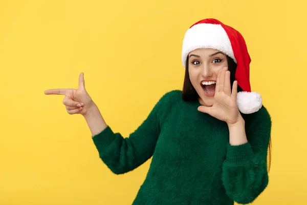 Fantastiska unga brunett kvinna bära Santa claus hatt och grön pullover peka finger bort över gul bakgrund. — Stockfoto