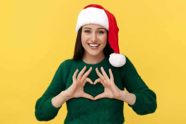 Glad ung brunett kvinna bära Santa claus hatt visar hjärta gest till kameran över gul bakgrund. — Stockfoto