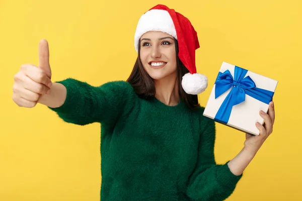 Feliz jovem morena usar chapéu de santa claus segurando caixa de presente e mostrar o polegar sobre fundo amarelo. — Fotografia de Stock