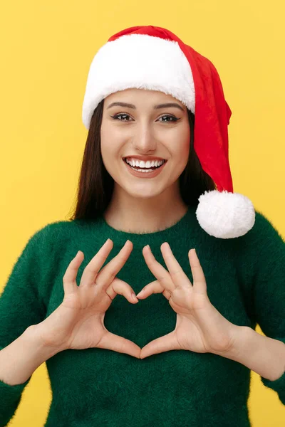 Charmig ung brunett kvinna bära Santa claus hatt visar hjärta gest till kameran över gul bakgrund. — Stockfoto