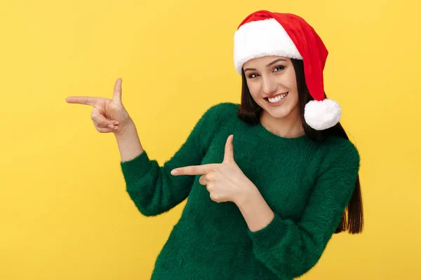 Mulher morena jovem atraente usar chapéu de Papai Noel e pulôver verde apontar dedos de distância sobre fundo amarelo. — Fotografia de Stock