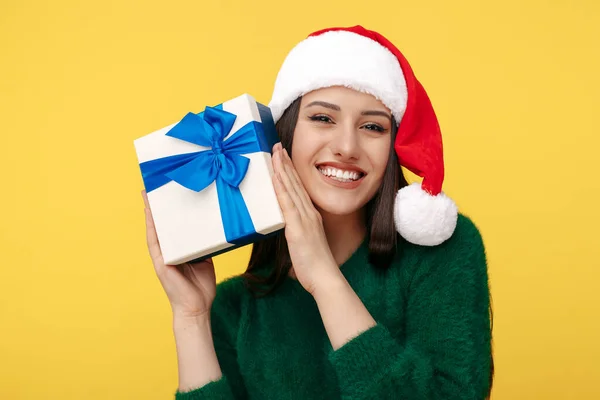 Feliz jovem morena usar chapéu de Papai Noel e pulôver verde segurando caixa de presente sobre fundo amarelo. — Fotografia de Stock