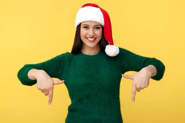 Encantadora jovem morena usar chapéu de Papai Noel e pulôver verde apontar dedos para baixo sobre fundo amarelo. — Fotografia de Stock