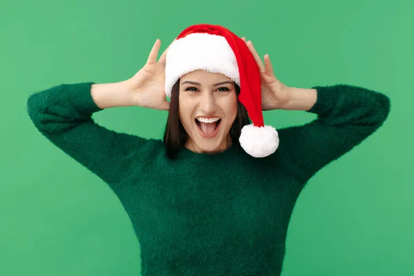 Spännande ung brunett kvinna bära Santa claus hatt och grön pullover över grön bakgrund. — Stockfoto