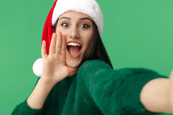 Skrik positiv ung brunett kvinna bära Santa claus hatt och grön pullover ta selfie över grön bakgrund. — Stockfoto