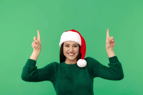 Happy young brunette woman wear santa claus hat and wool pullover point fingers up over green background. — Stock Photo, Image