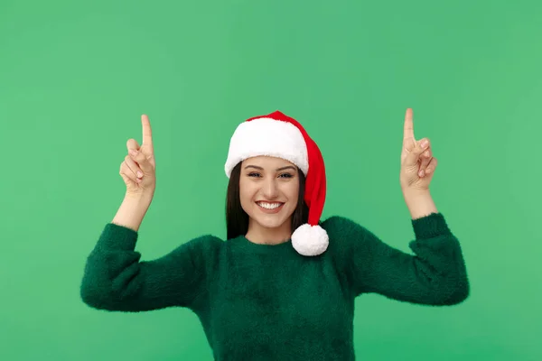 Feliz jovem morena mulher usar santa claus chapéu e lã pulôver apontar dedos para cima sobre fundo verde. — Fotografia de Stock