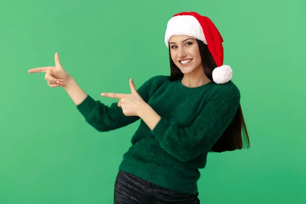 Cheerful young brunette woman wear santa claus hat and wool pullover point fingers awayover background. — Stock Photo, Image