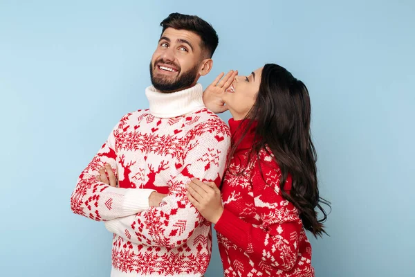 Mavi arka planda Noel süveteri giymiş neşeli bir çift. Bir kadın erkek kulağına fısıldıyor.
