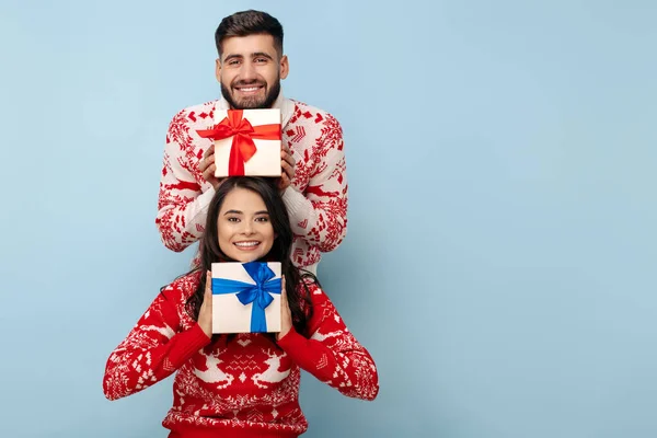 Casal alegre vestido com camisolas de Natal segurando caixas de presente sorrindo sobre fundo azul. — Fotografia de Stock