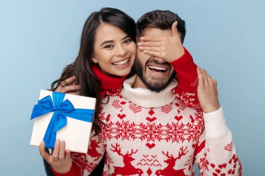 Noel süveteri giymiş neşeli bir çift. Kadın mavi arka planda erkek arkadaşının gözlerini kapatıyor..