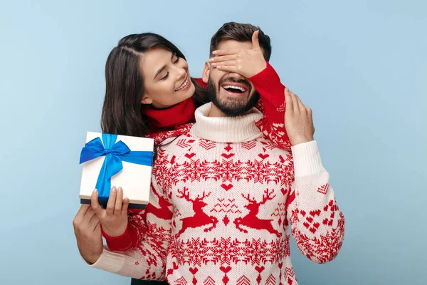 Atraente casal vestido com camisolas de Natal mulher cobrindo seus namorados olhos sobre fundo azul. — Fotografia de Stock