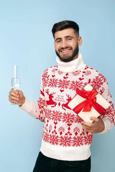 Bonito homem hipster vestido com camisola de Natal segurando caixa de presente e copo de champanhe sorrindo sobre fundo azul. — Fotografia de Stock