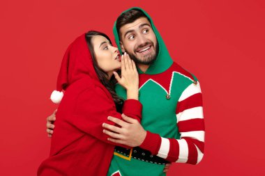 Kırmızı arka planda komik kazaklar giyen çekici bir çift. Bir kadın erkek kulağına fısıldıyor.