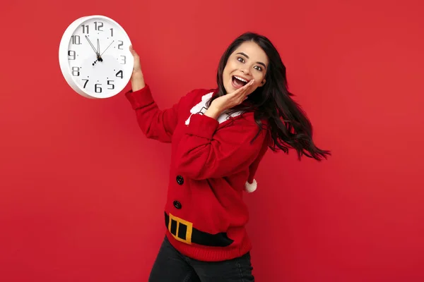 Surprise jeune femme brune habillée en pull Père Noël tenant horloge sur fond bleu. — Photo