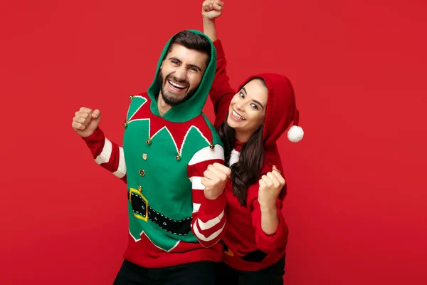 Spännande par klädda i jul roliga tröjor över röd bakgrund. Fira framgång. — Stockfoto