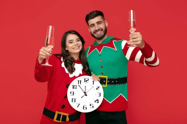Adorável casal em camisolas engraçadas de Natal segurando branco grande relógio e óculos com champanhe sobre fundo vermelho. — Fotografia de Stock