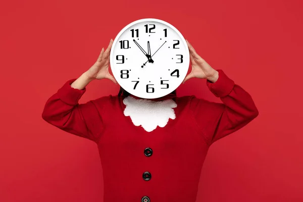 Junge brünette Frau im Weihnachtsmann-Pullover hält Uhr vor Gesicht vor blauem Hintergrund. — Stockfoto