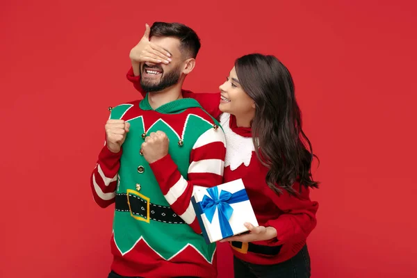 Atraente casal vestido com camisolas de Natal mulher cobrindo seus namorados olhos sobre fundo vermelho. — Fotografia de Stock